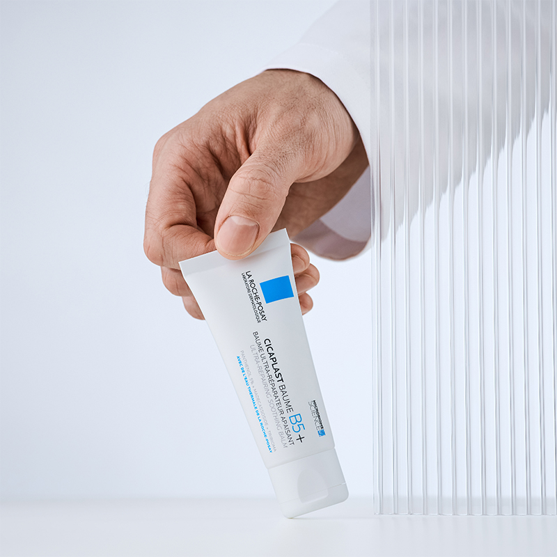 En hand håller en tub av La Roche-Posay Cicaplast Baume B5+ mot en ljus bakgrund. Tuben är vit med blå detaljer och text som beskriver produktens reparerande egenskaper. Bakgrunden har en transparent, ribbad struktur som ger en modern och ren känsla.