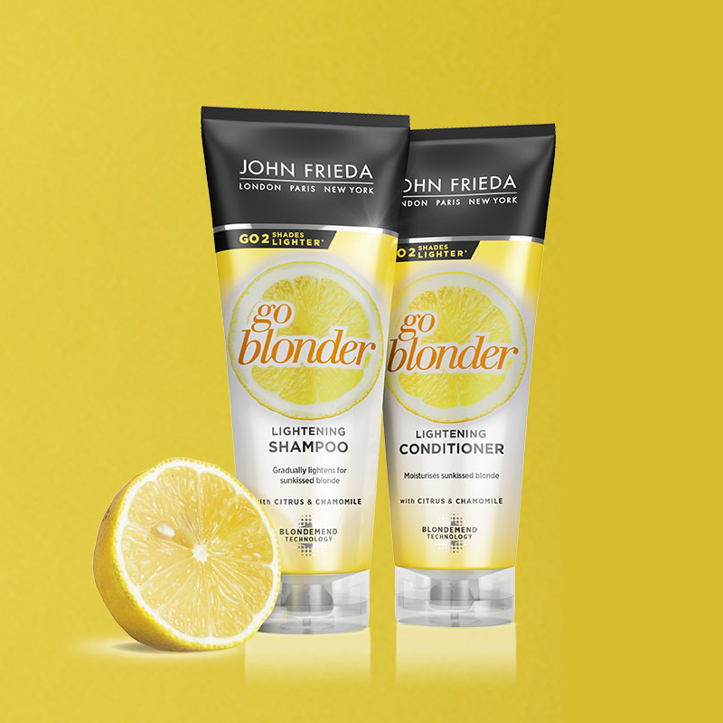 To hårpleieprodukter fra John Frieda, en sjampo og en balsam fra serien «Go Blonder». Produktene har et gult og hvitt design med et sitronmotiv på emballasjen. En halv sitron er plassert foran produktene mot en gul bakgrunn.