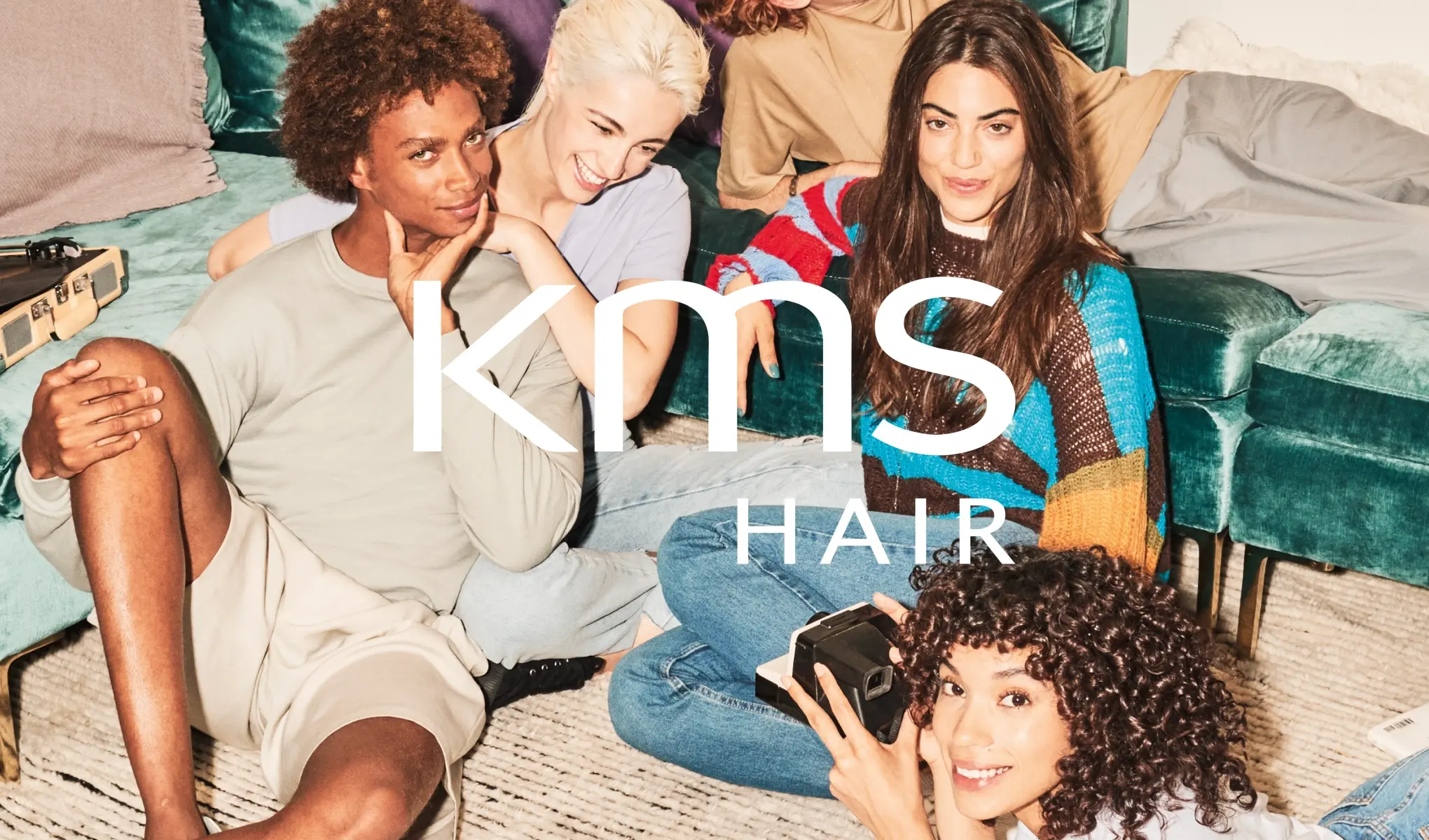 Useat henkilöt istuvat yhdessä olohuonemaisessa ympäristössä vihreiden sohvien edessä, ja kuvan päällä näkyy teksti “kms HAIR”. Yksi henkilö pitää kameraa kädessään, ja ryhmä esittelee erilaisia kampauksia ja hiustekstuureja KMS Hair -hiustenhoidon lifestyle-kuvassa.