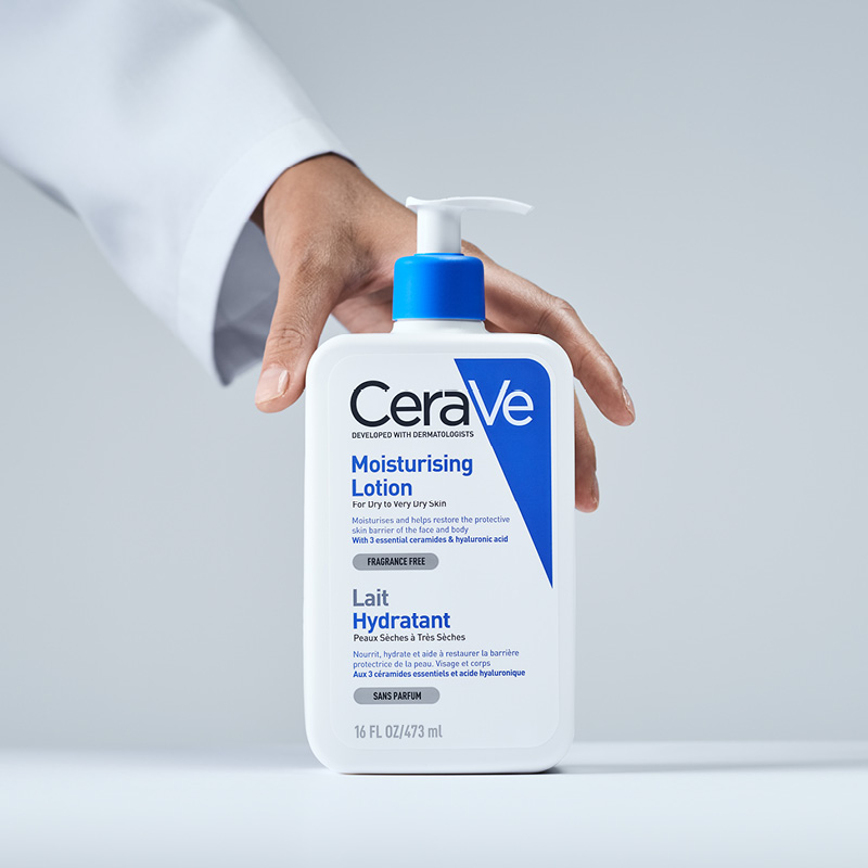 En hand som håller en flaska av CeraVes Moisturising Lotion. Flaskans etikett är vit med blå och svart text, och den har en blå pump. Bakgrunden är ljusgrå.