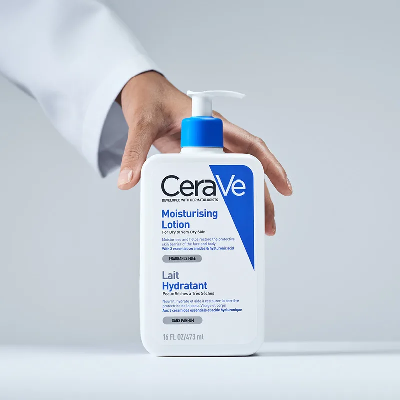En hand som håller en flaska av CeraVes Moisturising Lotion. Flaskans etikett är vit med blå och svart text, och den har en blå pump. Bakgrunden är ljusgrå.