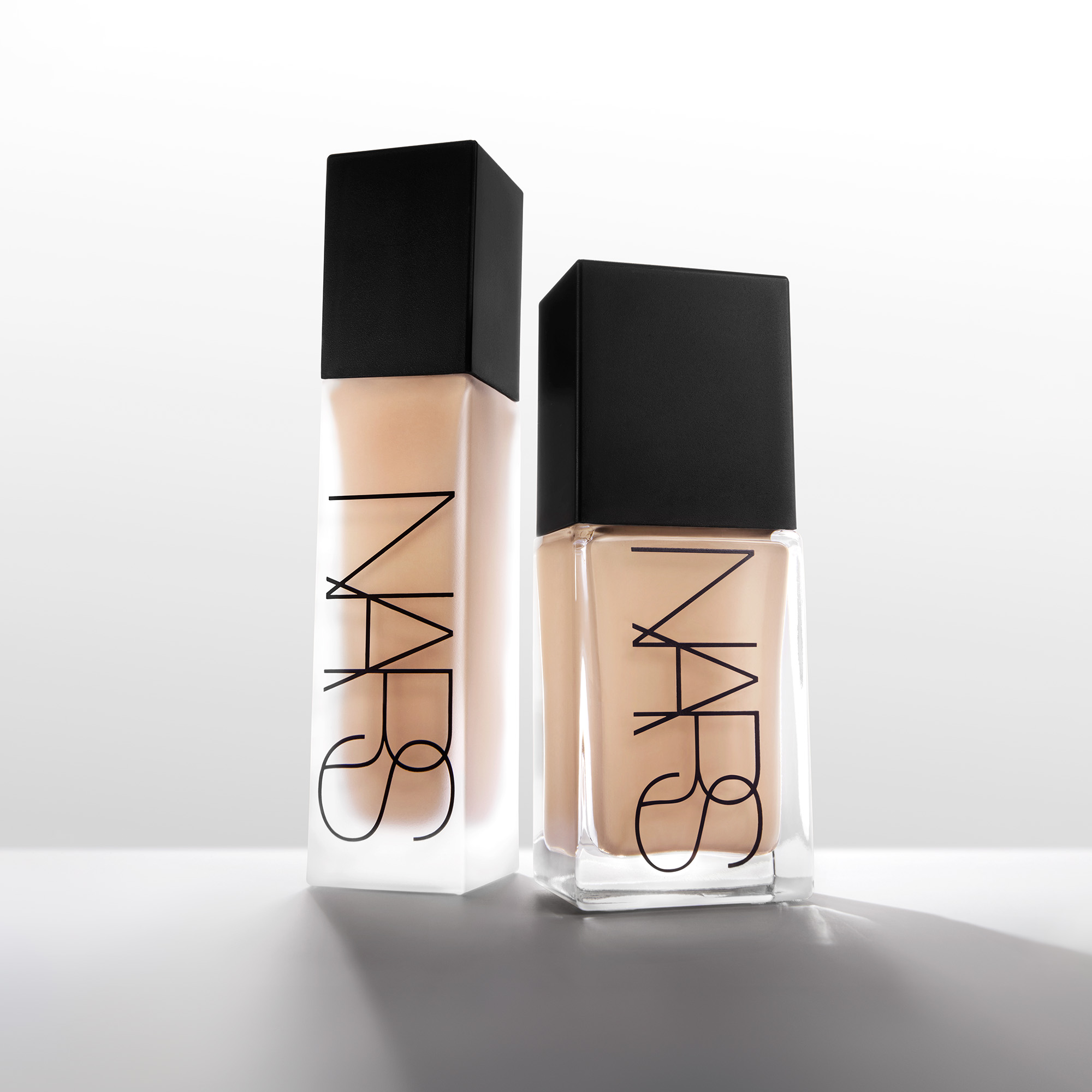 To NARS foundationflasker i lys beige nyanse står mot en lys bakgrunn, én med frostet finish og én i transparent glassflaske. Den svarte logoen “NARS” er tydelig synlig på begge emballasjene.