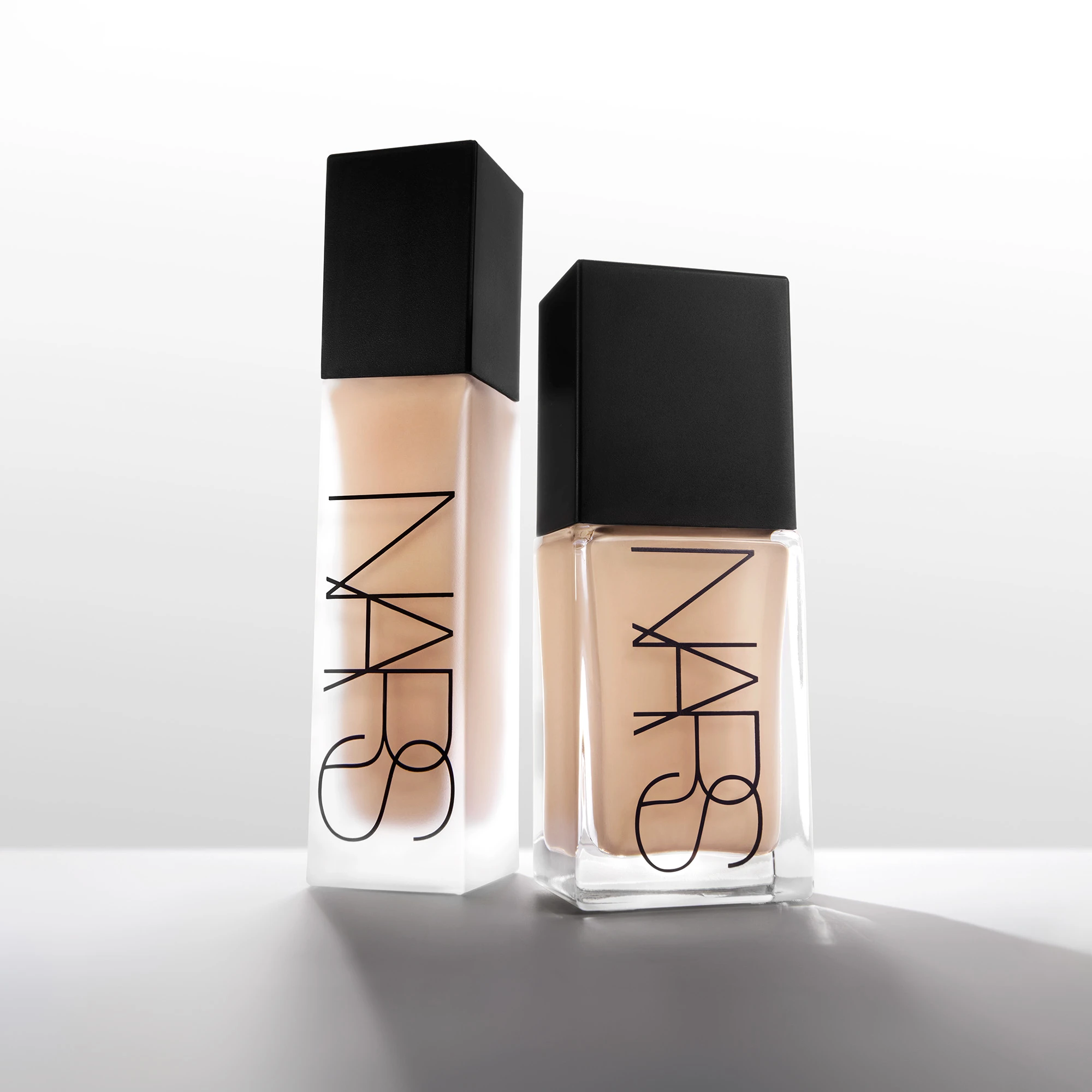To NARS foundationflasker i lys beige nyanse står mot en lys bakgrunn, én med frostet finish og én i transparent glassflaske. Den svarte logoen “NARS” er tydelig synlig på begge emballasjene.