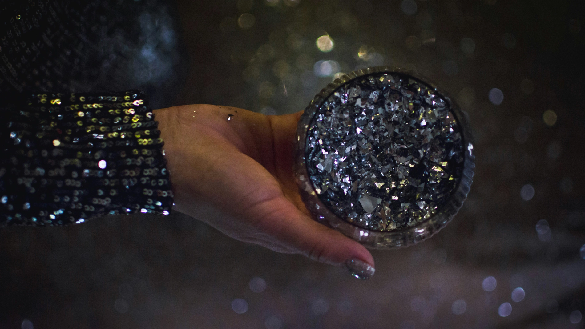 Käsi, jossa on lakatut kynnet ja tumma, kimaltava hiha, pitelee pyöreää, tummalla glitterillä päällystettyä rasiaa hämärää ja sumeaa taustaa vasten.