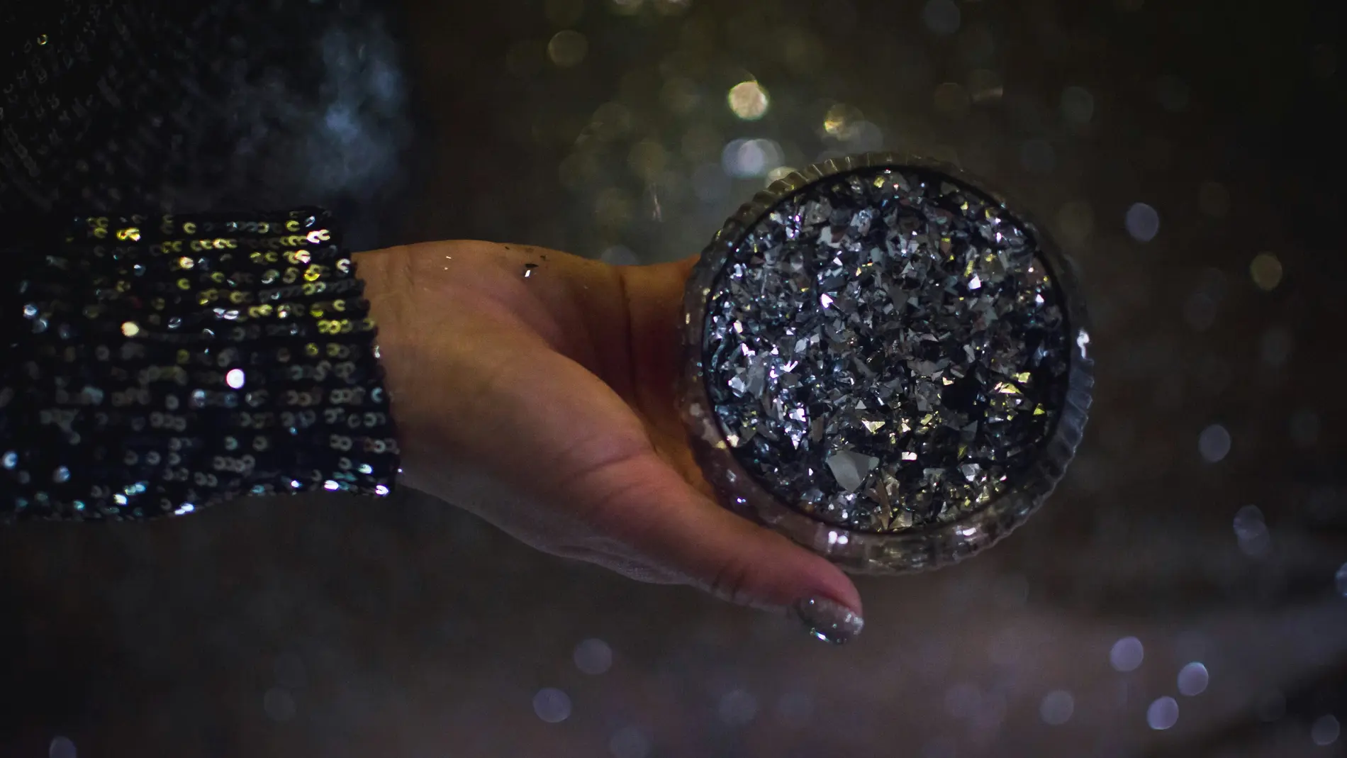 Käsi, jossa on lakatut kynnet ja tumma, kimaltava hiha, pitelee pyöreää, tummalla glitterillä päällystettyä rasiaa hämärää ja sumeaa taustaa vasten.