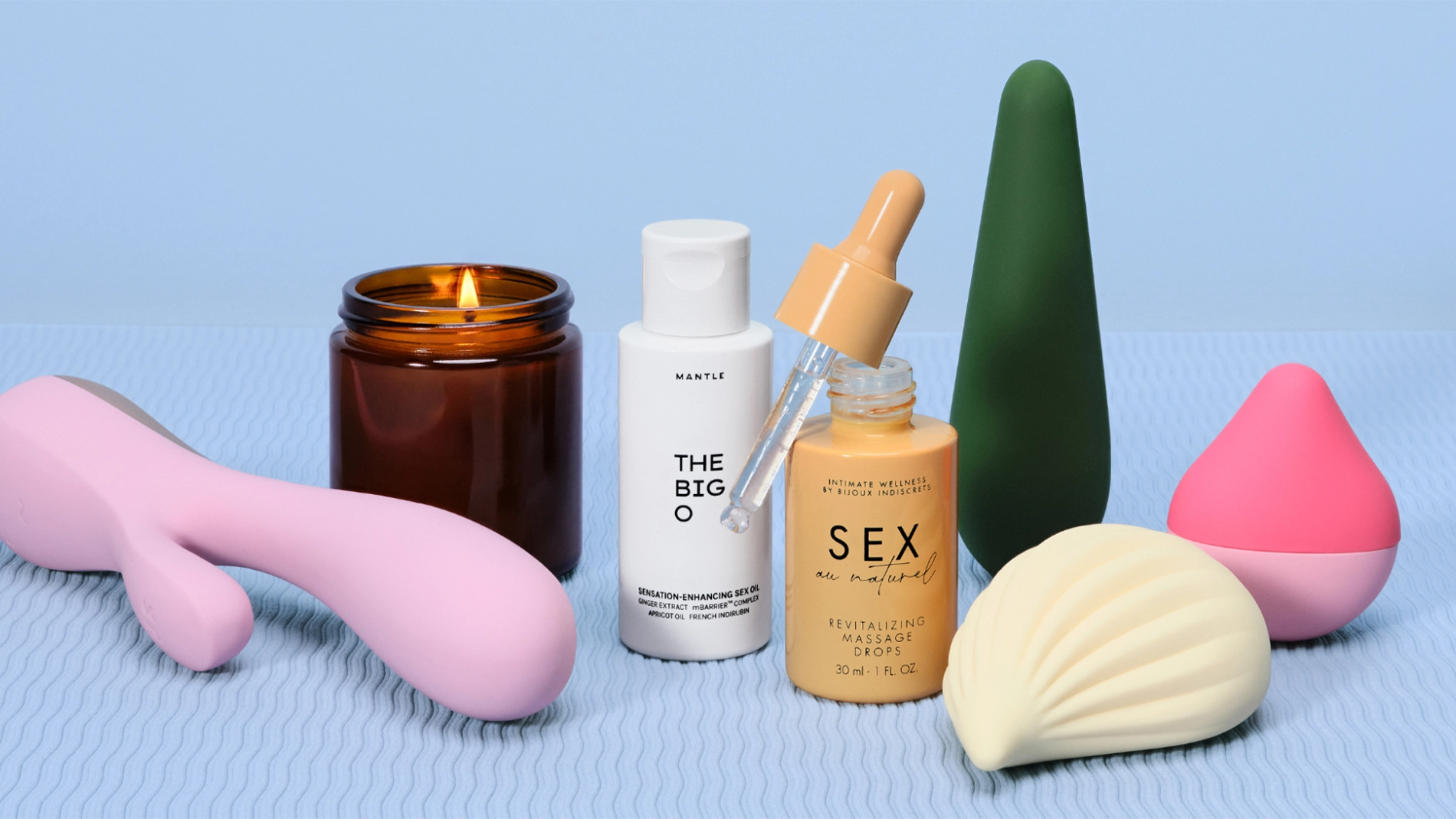 Stillbild med intimprodukter och kroppsvård på ljusblå bakgrund. Ett tänt doftljus står bredvid Mantle The Big S*xcation Enhancing Serum och en flaska “Sex Oil” revitalizing massage oil med pipett, tillsammans med rosa och gröna sexleksaker.