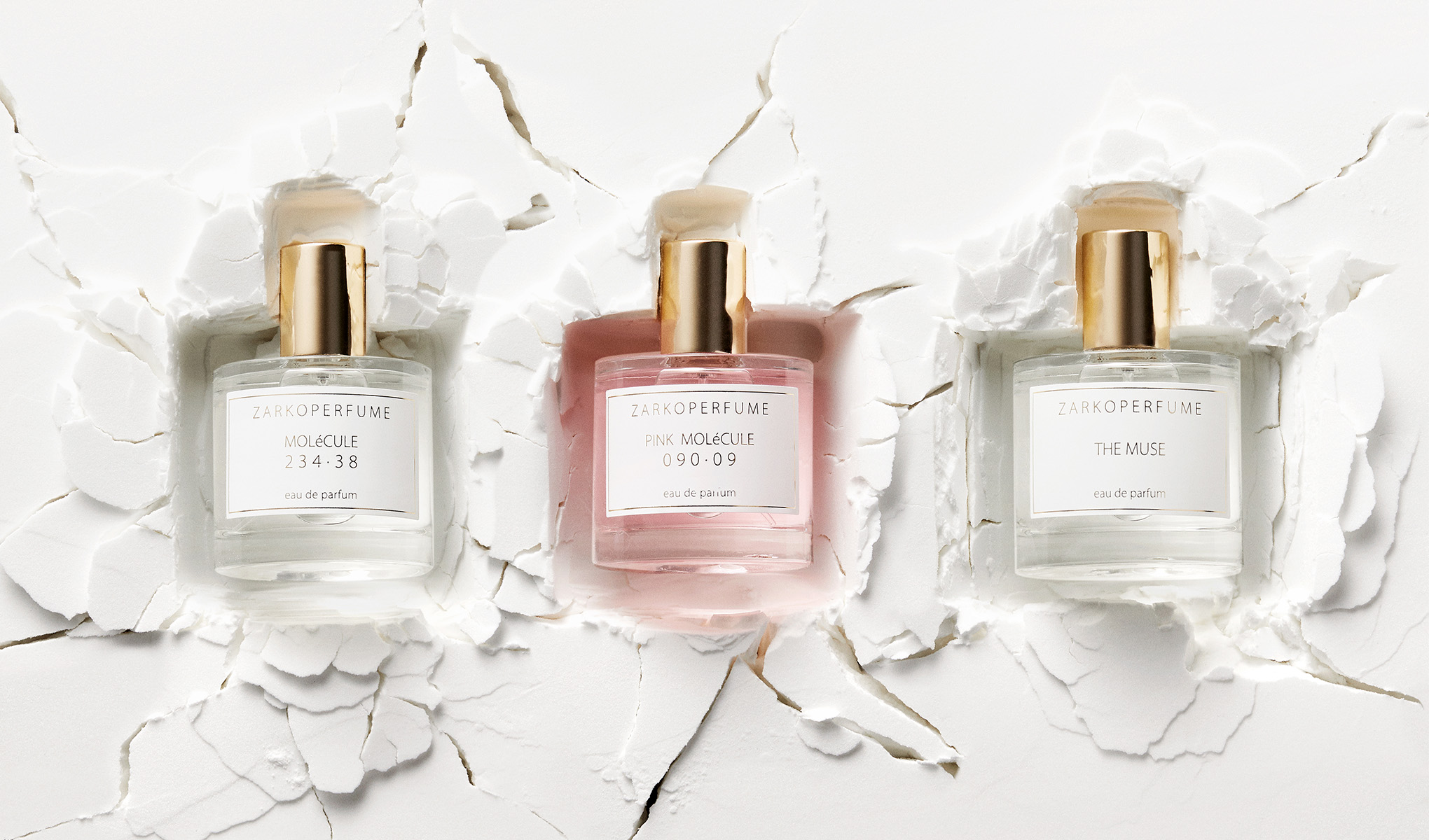 Tre Zarkoperfume-flasker på en hvit, sprukket bakgrunn. Til venstre Molécule, i midten Pink Molécule og til høyre The Muse. Flaskene har gullfargede korker.