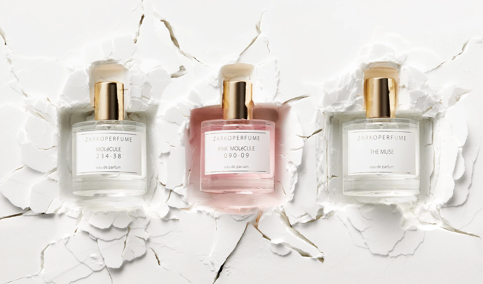 Tre Zarkoperfume-flasker på en hvit, sprukket bakgrunn. Til venstre Molécule, i midten Pink Molécule og til høyre The Muse. Flaskene har gullfargede korker.