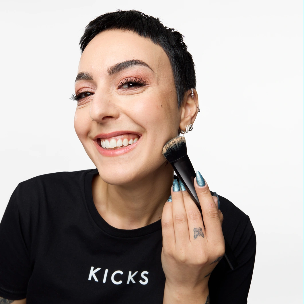 Person med kort svart hår påfører makeup med sminkekost mot kinnet og har på seg en svart KICKS-t-skjorte mot lys grå bakgrunn. Synlige detaljer inkluderer sølvfargede øredobber og blå neglelakk.