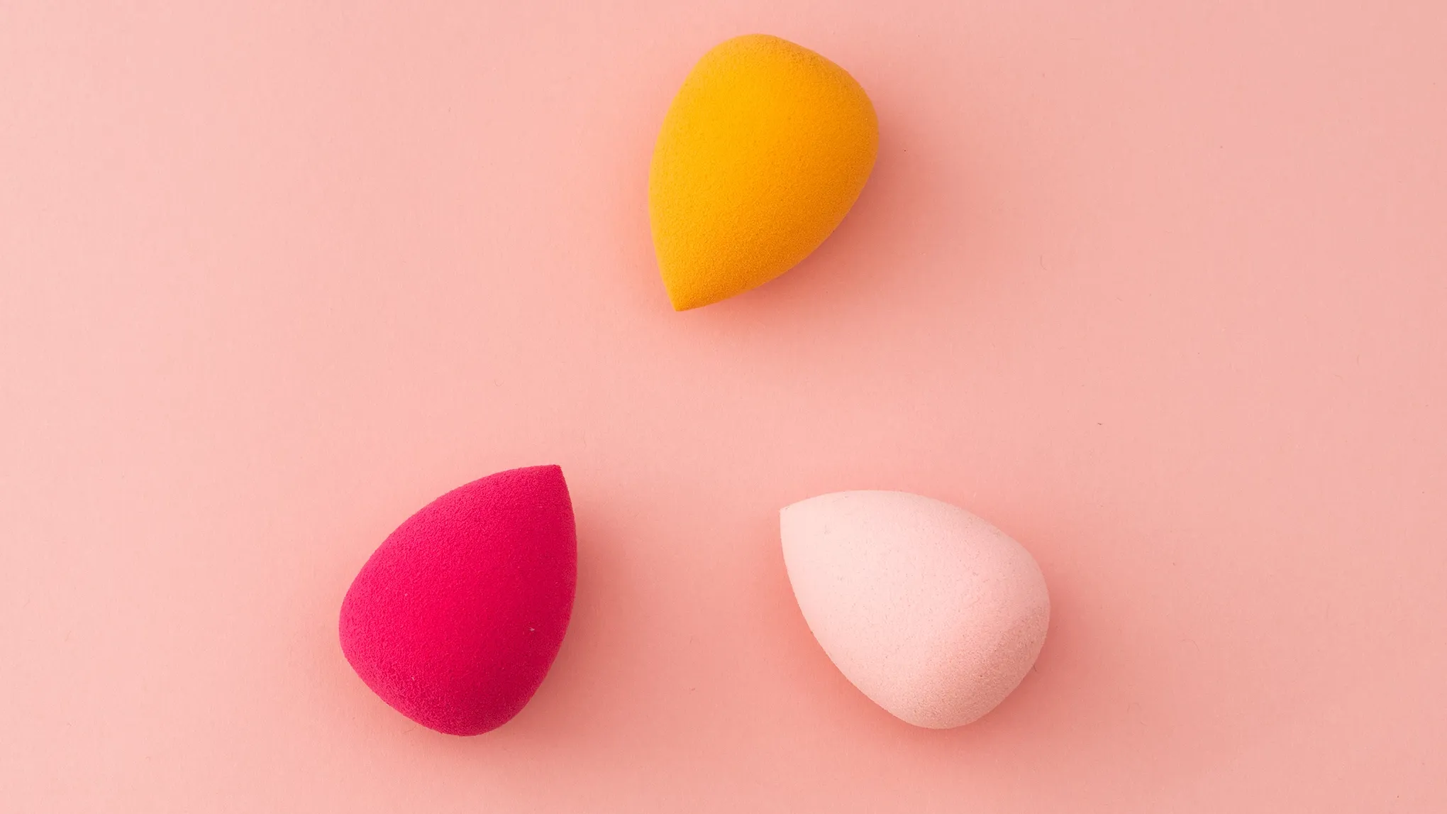 Tre sminksvamper i forskjellige farger (gul, rosa og rød) på en rosa bakgrunn.