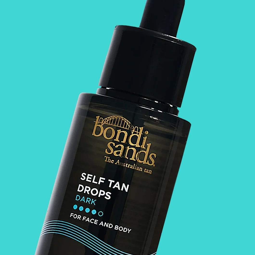 Bondi Sands Self Tan Drops Dark i en mørk glassflaske med pipette mot turkis bakgrunn. På emballasjen står tekstene «Bondi Sands», «The Australian Tan», «SELF TAN DROPS», «DARK» og «FOR FACE AND BODY».