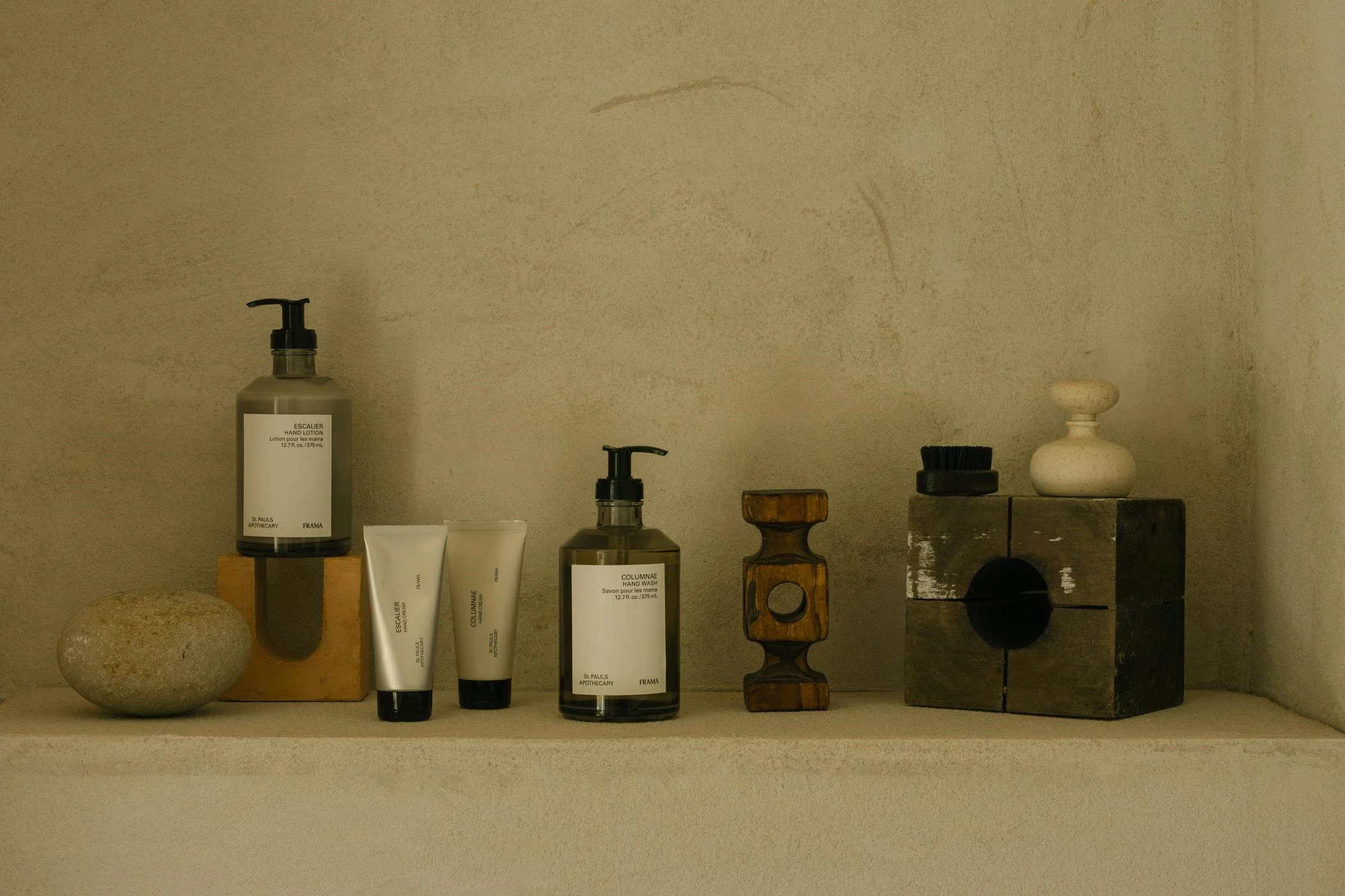 Två flaskor med handtvål och handlotion samt två tuber med hudvårdsprodukter står på en hylla tillsammans med dekorativa träföremål och en sten. Bakgrunden är en ljus, texturerad vägg.