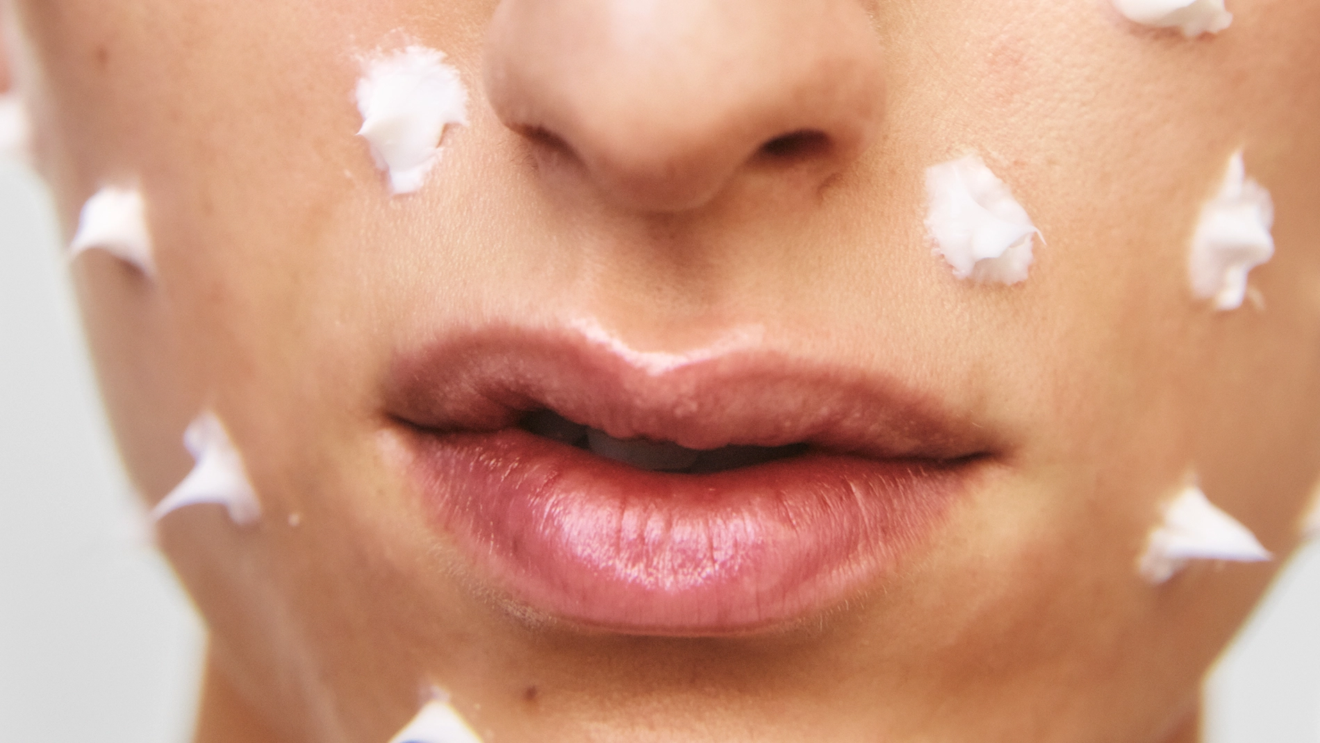 Nærbilde av ansikt med små klatter hvit ansiktskrem jevnt påført over kinnene og rundt munnen. Fokus på hudpleie og fuktighetskrem før den smøres inn.