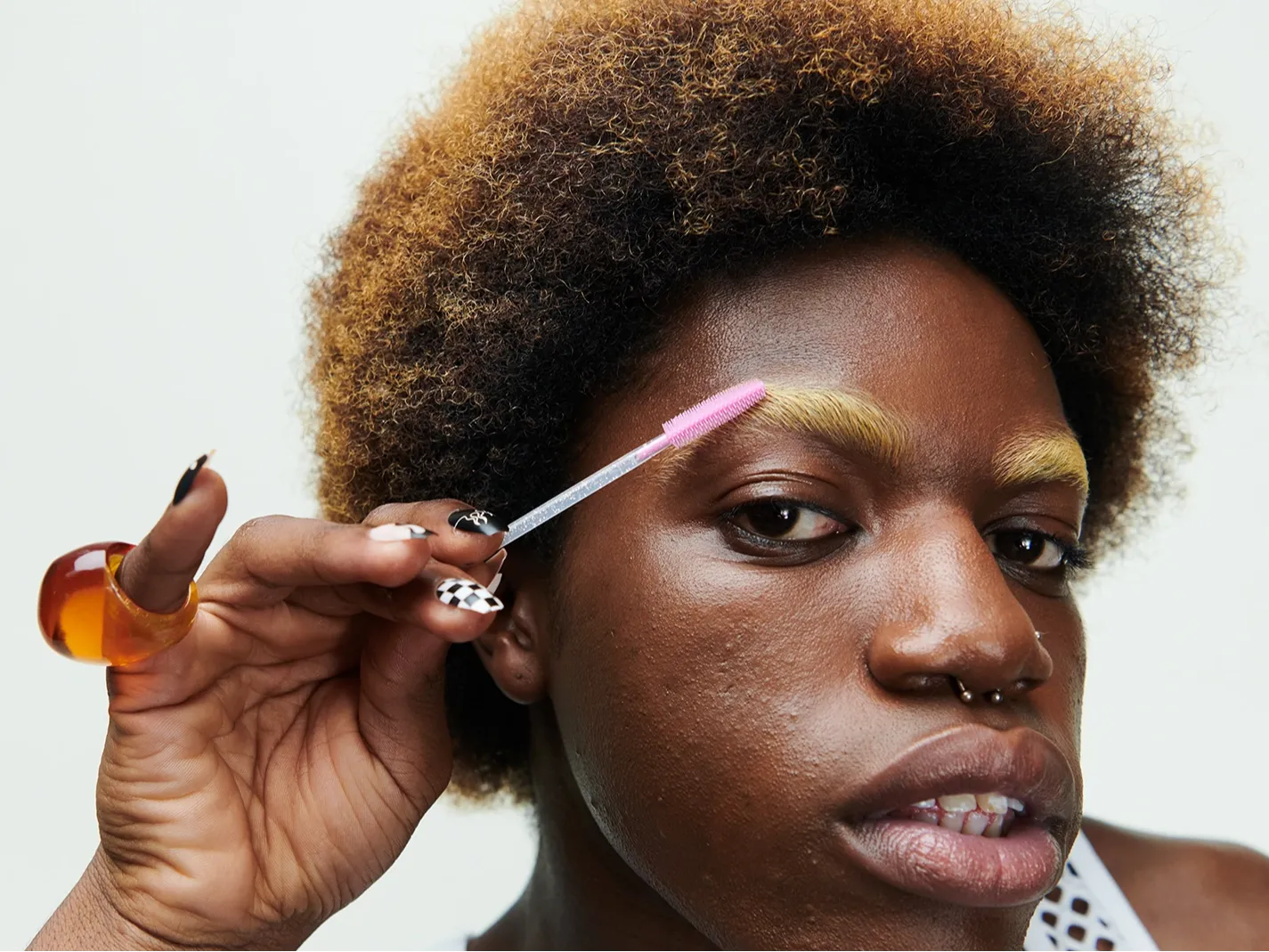 Nærbilde av en person som påfører brynsgel med en rosa spooliebørste på blekede øyenbryn, med fokus på brynsminke og hudtekstur. Naturlig makeup-look med definerte bryn og nøytral bakgrunn.