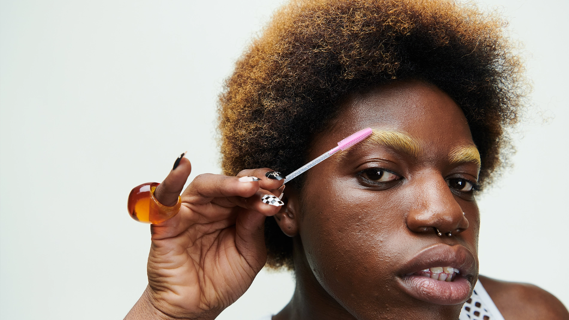 Lähikuva henkilöstä levittämässä kulmageeliä vaaleiksi käsiteltyihin kulmakarvoihin vaaleanpunaisella spoolie-harjalla, korostaen kulmameikkiä ja ihon tekstuuria. Luonnollinen meikkilook, jossa korostetut kulmat ja neutraali tausta.