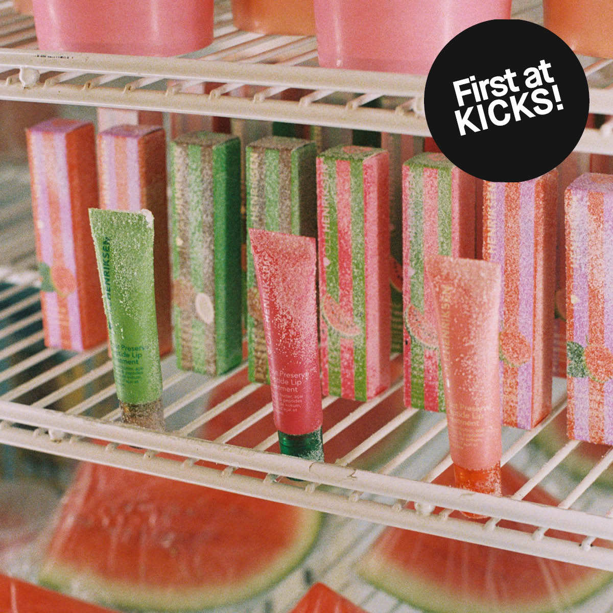 Fargerike Ole Henriksen leppeprodukter og esker i grønt, rosa og oransje står på en frostet hylle, med teksten “Ole Henriksen” og “Lip Treatment” synlig på tubene. En svart sirkel med teksten “First at KICKS!” vises over bildet, og vannmelon er synlig i bakgrunnen.