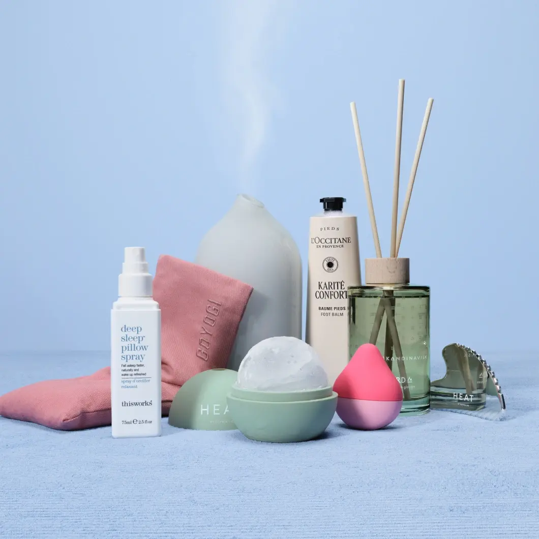 Wellnessprodukter arrangerade på ljusblå bakgrund med doftdiffuser, doftpinnar, fotkräm, pillow spray, rosa strumpor och beautyverktyg. Produkter från bland annat thisworks, L’OCCITANE, SKANDINAVISK och HEAT syns tillsammans med en rund bath bomb och en silverfärgad gua sha.