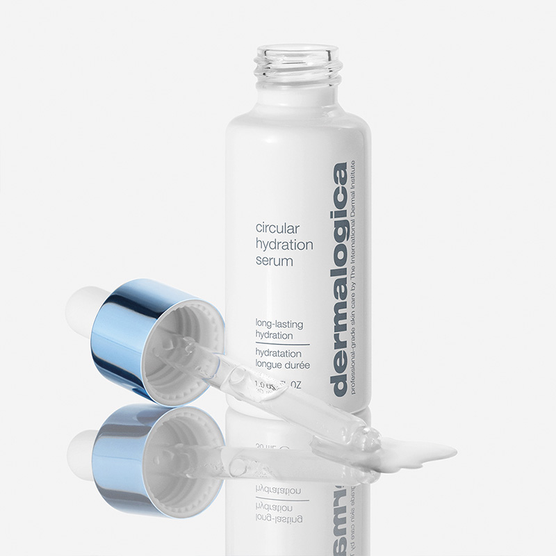 En flaska "Circular Hydration Serum" från märket Dermalogica. Flaskan är vit med texten "dermalogica" tryckt vertikalt längs sidan. Locket, som är blått med en pipett, ligger bredvid flaskan. En liten mängd serum har droppats ut från pipetten. Produkterna står på en reflekterande yta som skapar en spegeleffekt. Bakgrunden är ljus och neutral.