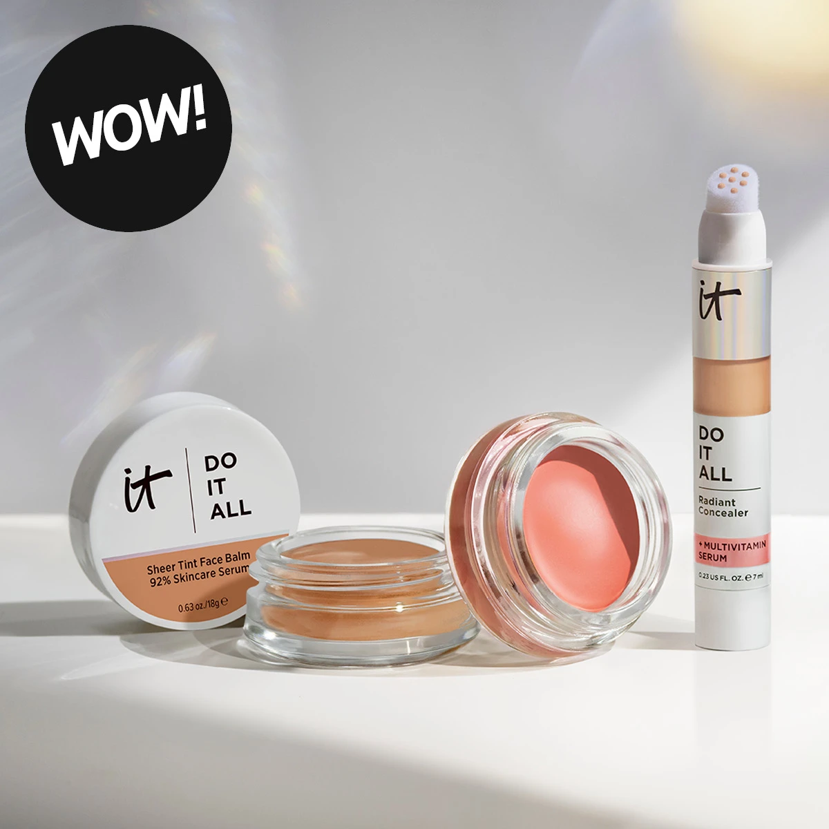 WOW! IT Cosmetics Do It All Sheer Tint Face Balm 92% Skincare Serum 0.63 oz/18 g og Do It All Radiant Concealer + Multivitamin Serum 0.23 US fl. oz./7 ml vises sammen med åpne makeupkrukker i beige og rosa toner.
