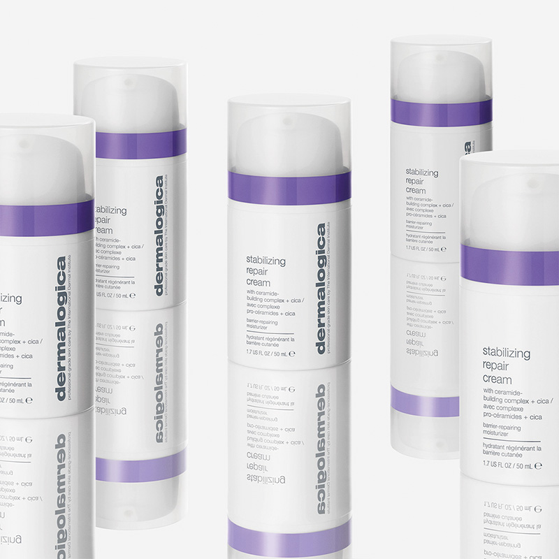 Flera flaskor av produkten "Stabilizing Repair Cream" från märket Dermalogica. Flaskorna är vita med lila detaljer och har texten "dermalogica" tryckt vertikalt längs sidan. Produkterna står på en reflekterande yta som skapar en spegeleffekt. Bakgrunden är ljus och neutral.