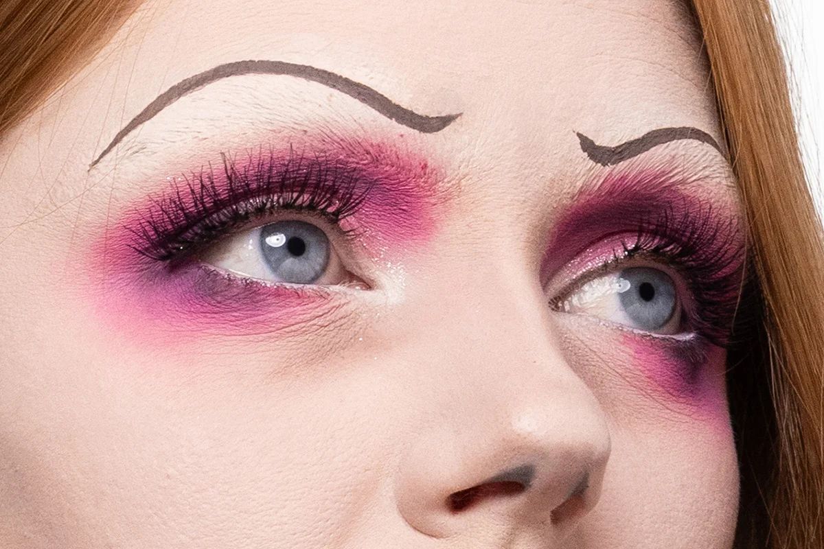 Närbild av ögonmakeup i starka rosa och lila skuggor med långa fransar och målade tunna, böjda ögonbryn som förstärker en karaktäristisk makeupstil.