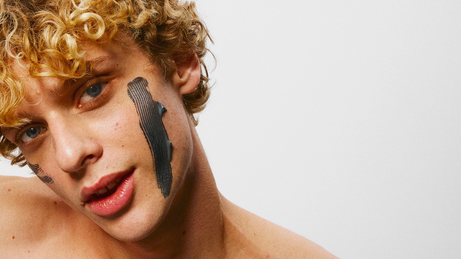 Närbild på person med lockigt blont hår och svart ansiktsmask applicerad i breda drag på kinderna.