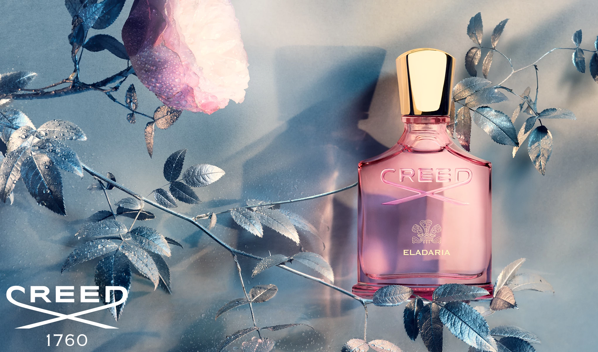 En rosa parfymflaska med guldkork och etiketten ”Creed Eladaria” står på en yta omgiven av silverfärgade blad och en rosa blomma. Bakgrunden är ljus och skapar en elegant och drömsk atmosfär. Creeds logotyp och årtalet 1760 syns längst ned till vänster i bilden.