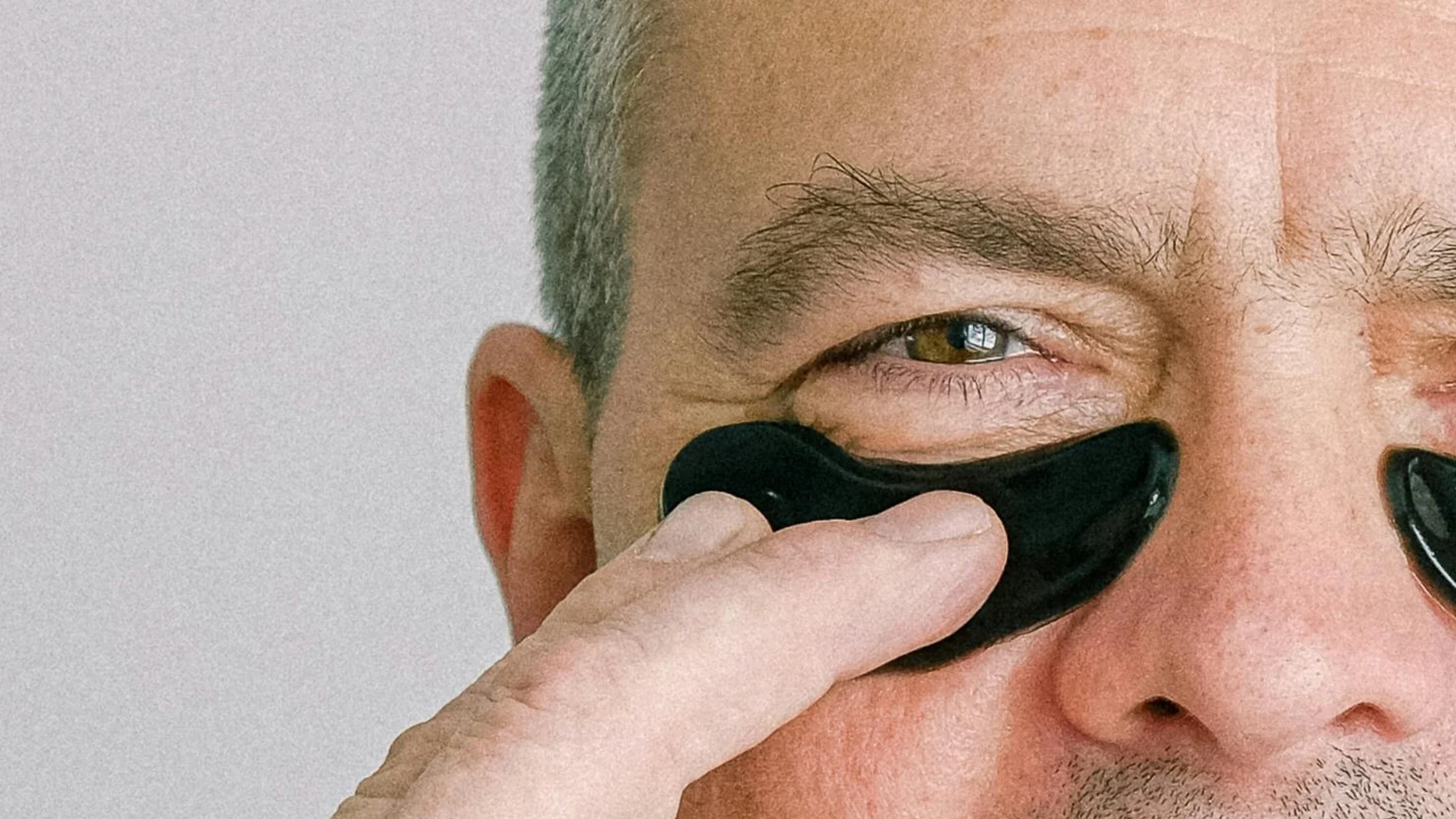 Närbild på en person med en svart ögonmask under ögat som han håller fast med ett finger. Personen är gråhårig och bakgrunden är ljusgrå.