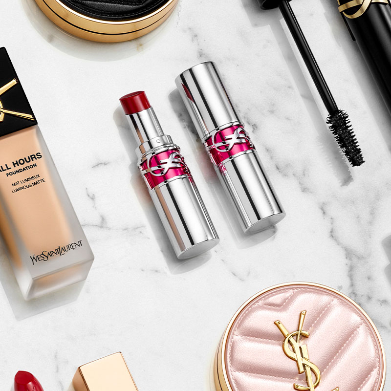 Flera sminkprodukter från Yves Saint Laurent, arrangerade på en vit marmorbakgrund. I mitten finns två silverfärgade läppstift, varav ett är öppet och visar en röd färg. Runt dem finns en foundationflaska, en mascara, en puderkompakt med YSL-logotypen, och en röd läppstiftshylsa med guldkork. Alla produkter har eleganta förpackningar med YSL-logotypen.