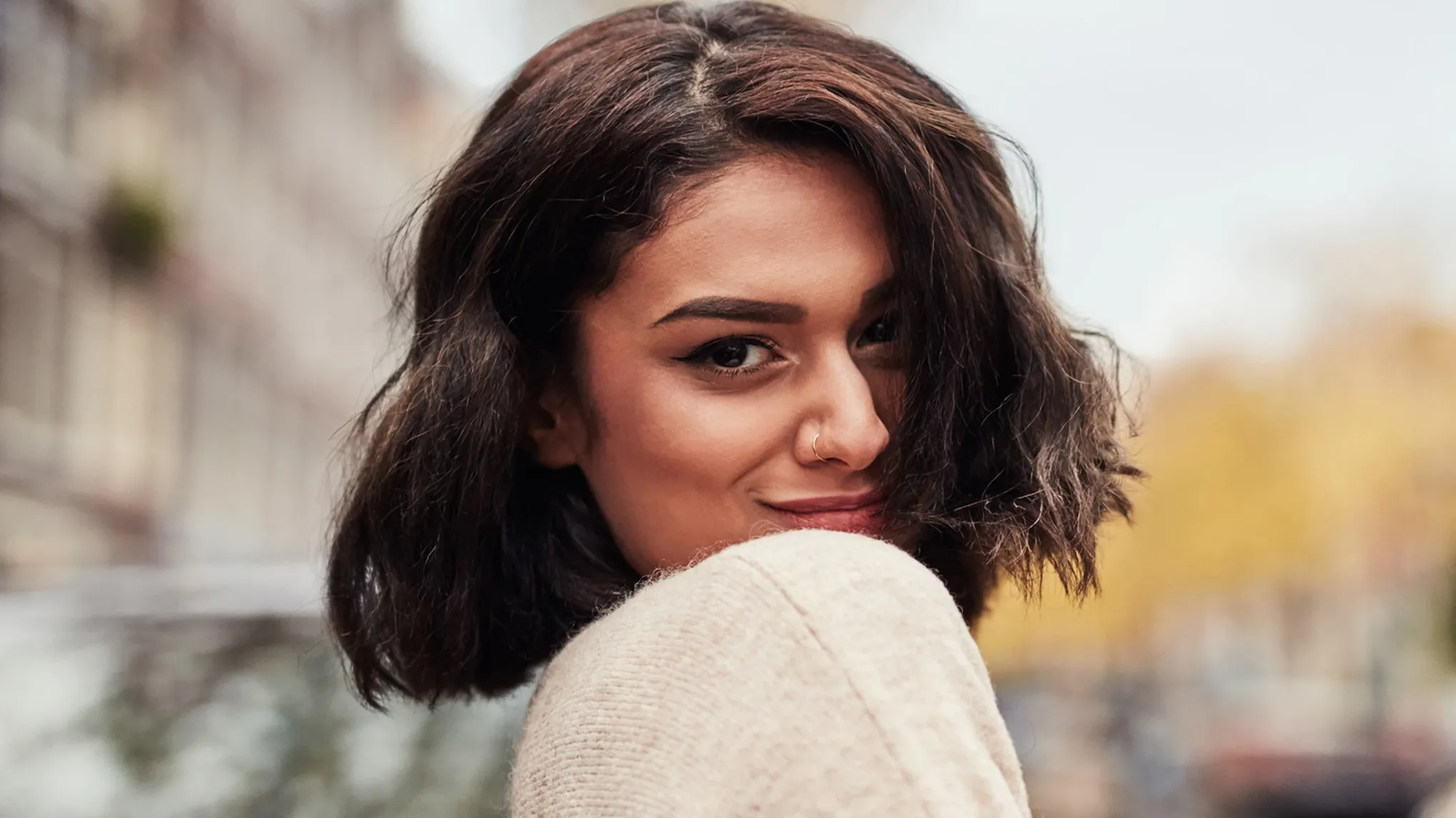 Kvinna med kort, rufsig, pagefrisyr som ler över sin axel med suddig stadsmiljö i bakgrunden.