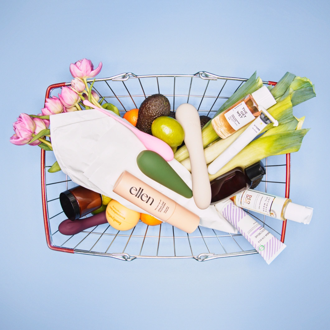 En kundvagn fylld med wellness- och intimvårdsprodukter visas mot ljusblå bakgrund. Kundvagnen innehåller bland annat blommor, avokado, citrusfrukter, sexleksaker och produkter från ellen, DeoDoc och The Get Wet.