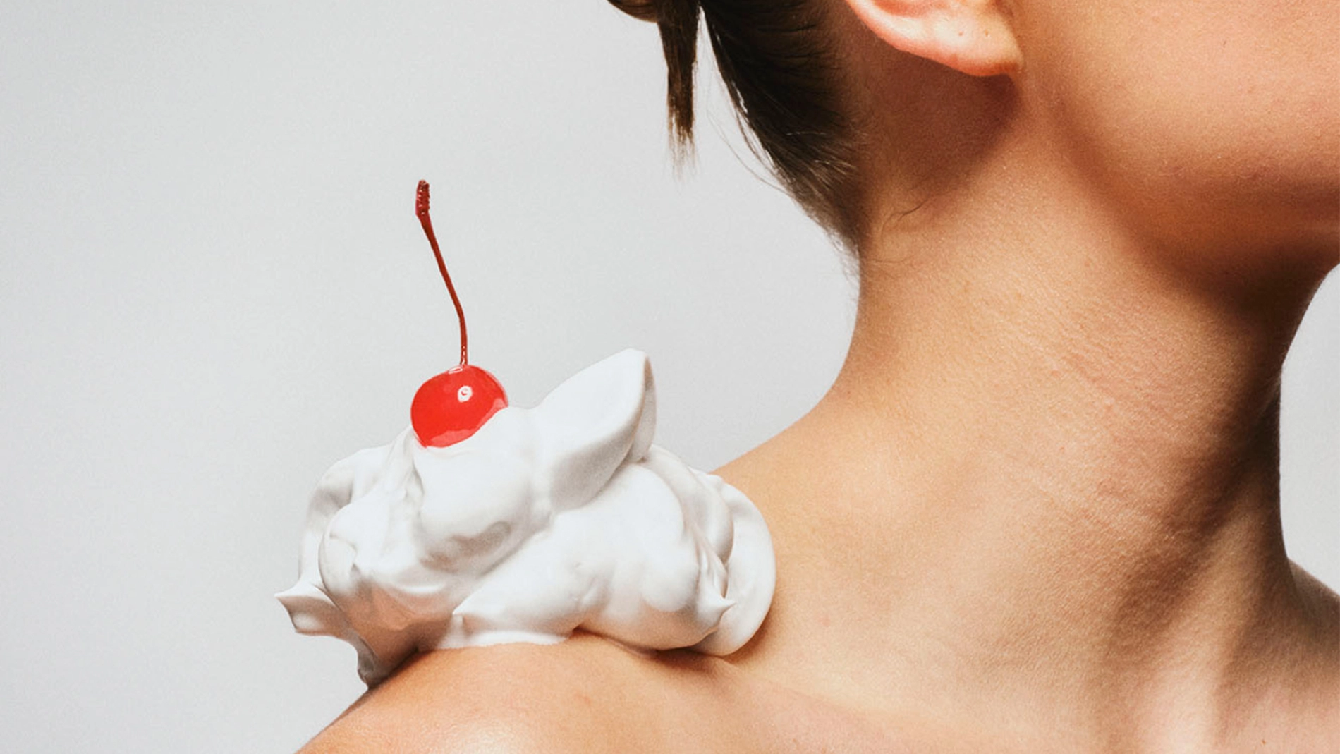 Nærbilde av en bar skulder med en klatt hvit krem plassert på huden, toppet med et rødt kirsebær, mot nøytral bakgrunn.