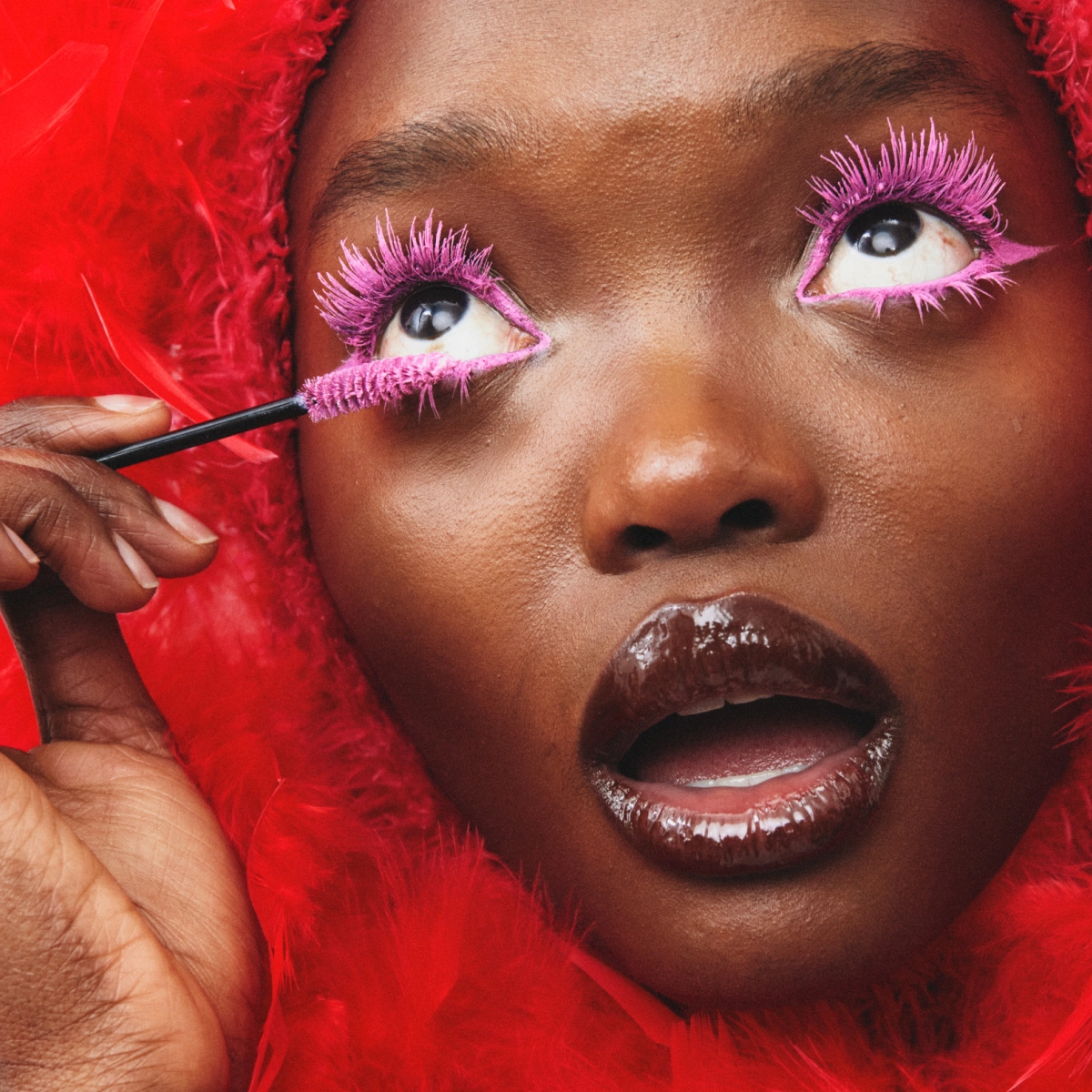 Lähikuva henkilöstä levittämässä pinkkiä ripsiväriä alaripsiin, pinkki eyeliner ja kiiltävät huulet sekä punainen pörröinen tausta.