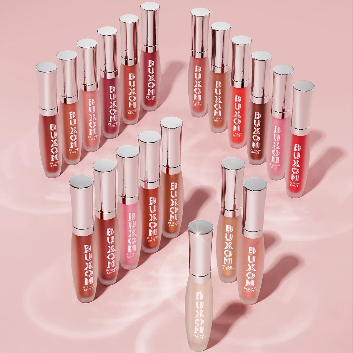 En samling av Buxom Plump Shot lipgloss i forskjellige farger, arrangert på en rosa bakgrunn.