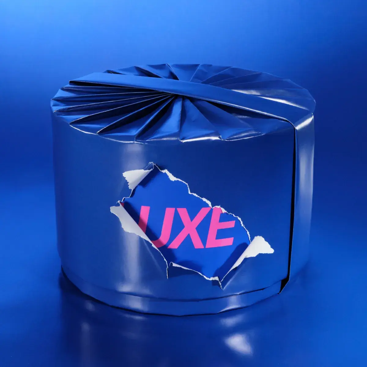 Ett runt paket inslaget i glänsande blått papper mot en blå bakgrund. Rosa ”LUXE”-text syns genom en riven öppning.