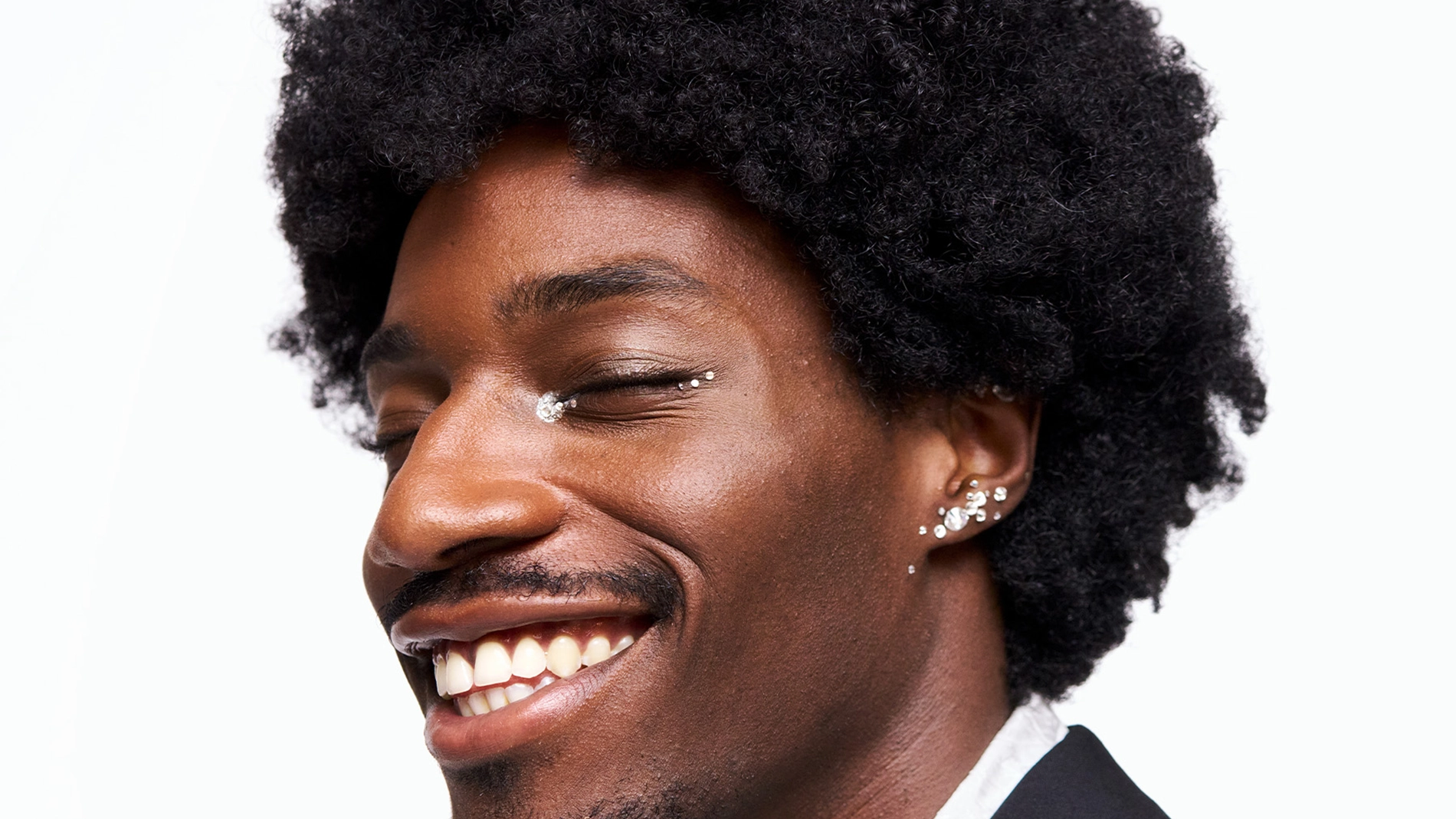 Kuva, joka näyttää henkilön, jolla on mustat kiharat hiukset ja useita korvakoruja korvassa. Henkilöllä on yllään musta takki ja valkoinen paita. Tausta on valkoinen.