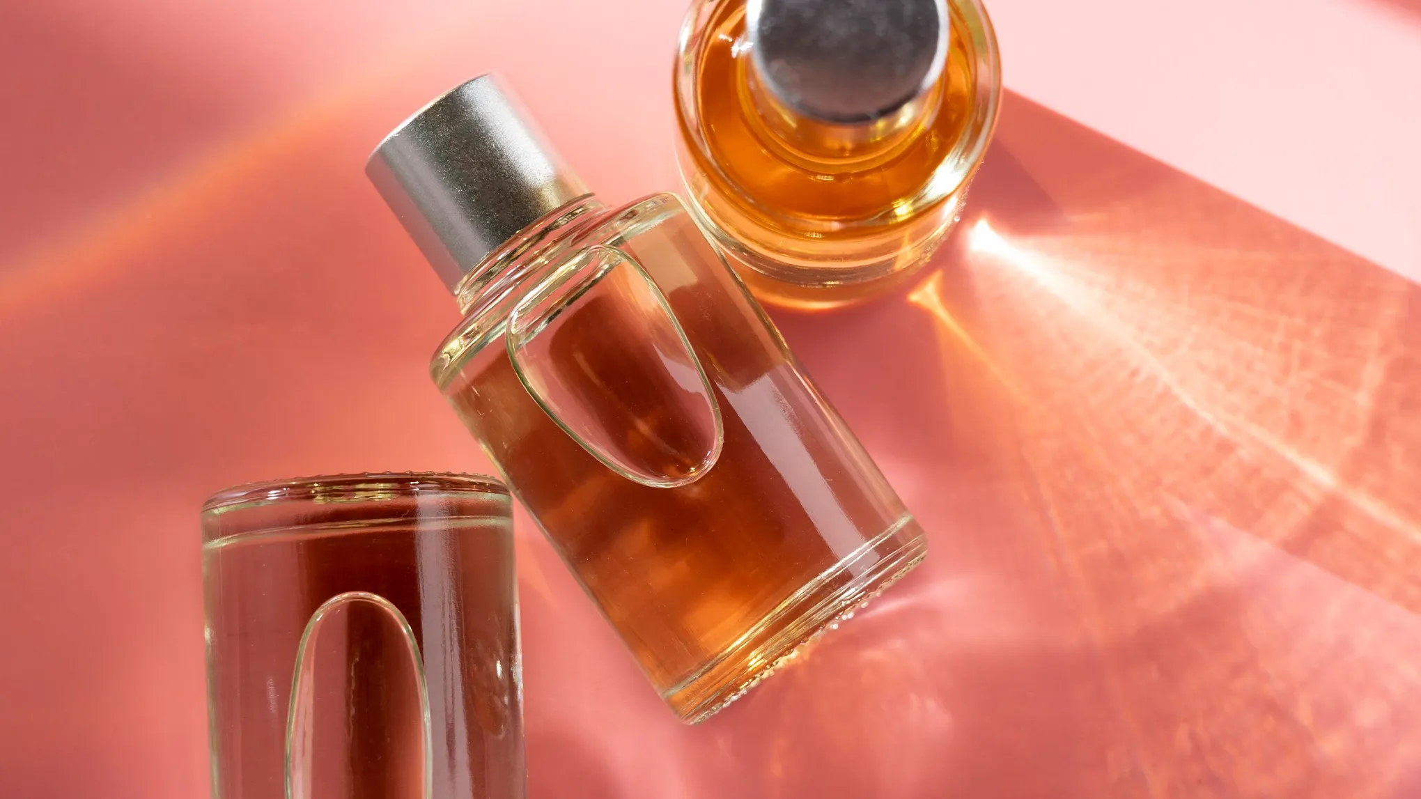 Tre parfymflaskor med orange vätska på en rosa bakgrund.