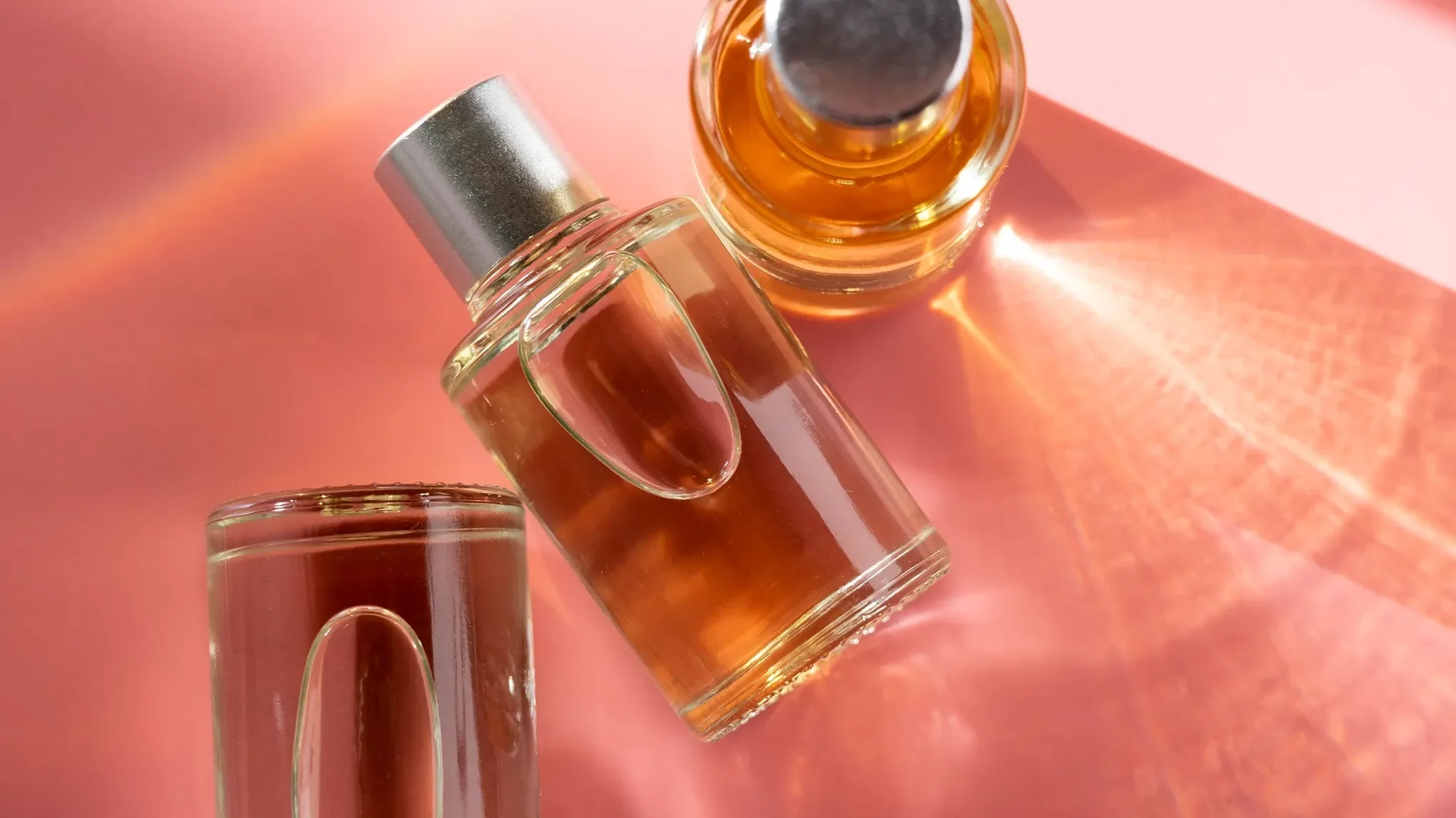 Tre parfymeflasker med oransje væske på en rosa bakgrunn.