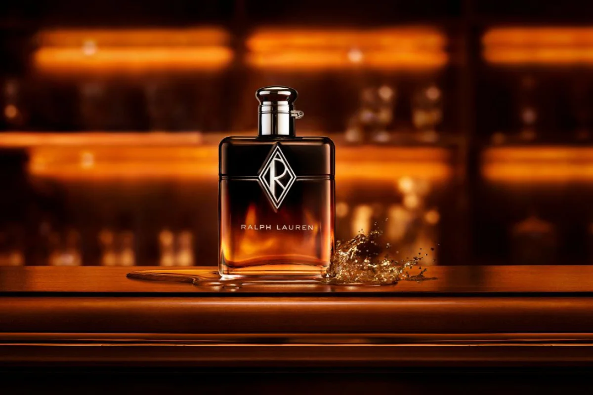 Ralph Lauren-parfymeflaske med synlig tekst «RALPH LAUREN» og monogram plassert på en treflate foran en varmtonet, uskarp bakgrunn. En væskesprutdetalj vises ved siden av flasken.