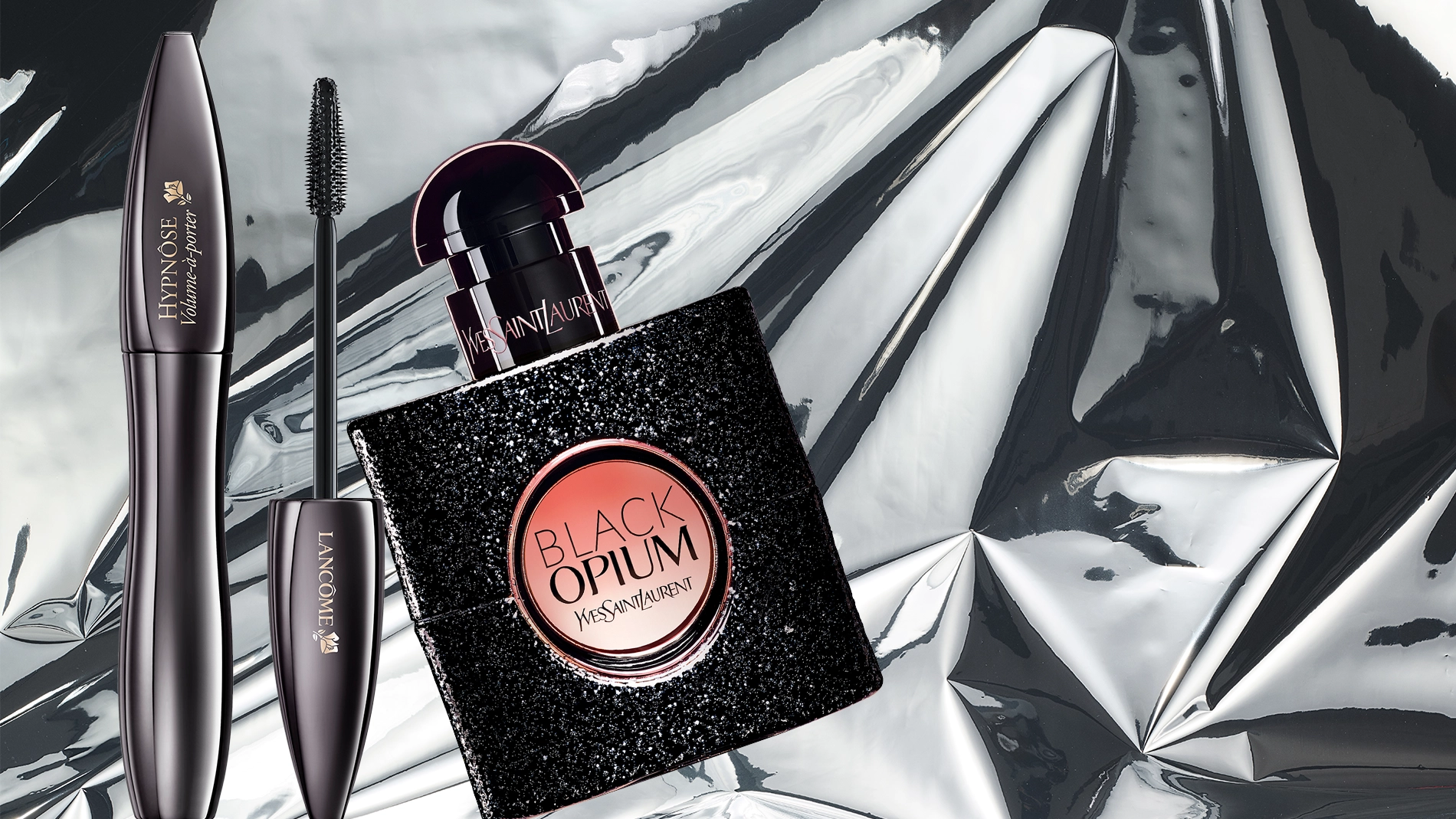 Lancôme Hypnôse Volume-maskara og Yves Saint Laurent Black Opium-parfyme er plassert mot en blank sølvbakgrunn som fremhever en glamorøs beautystemning.