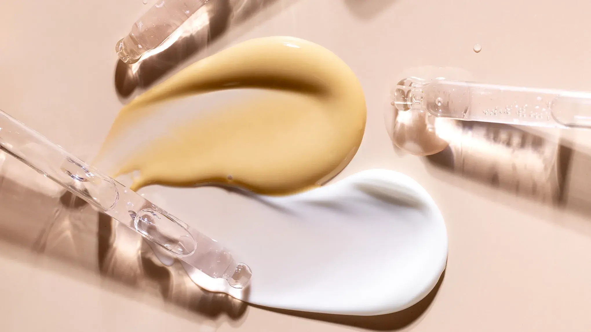 Nærbilde av to forskjellige hudpleieprodukter, en gul og en hvit, påført på en beige overflate med pipetter.