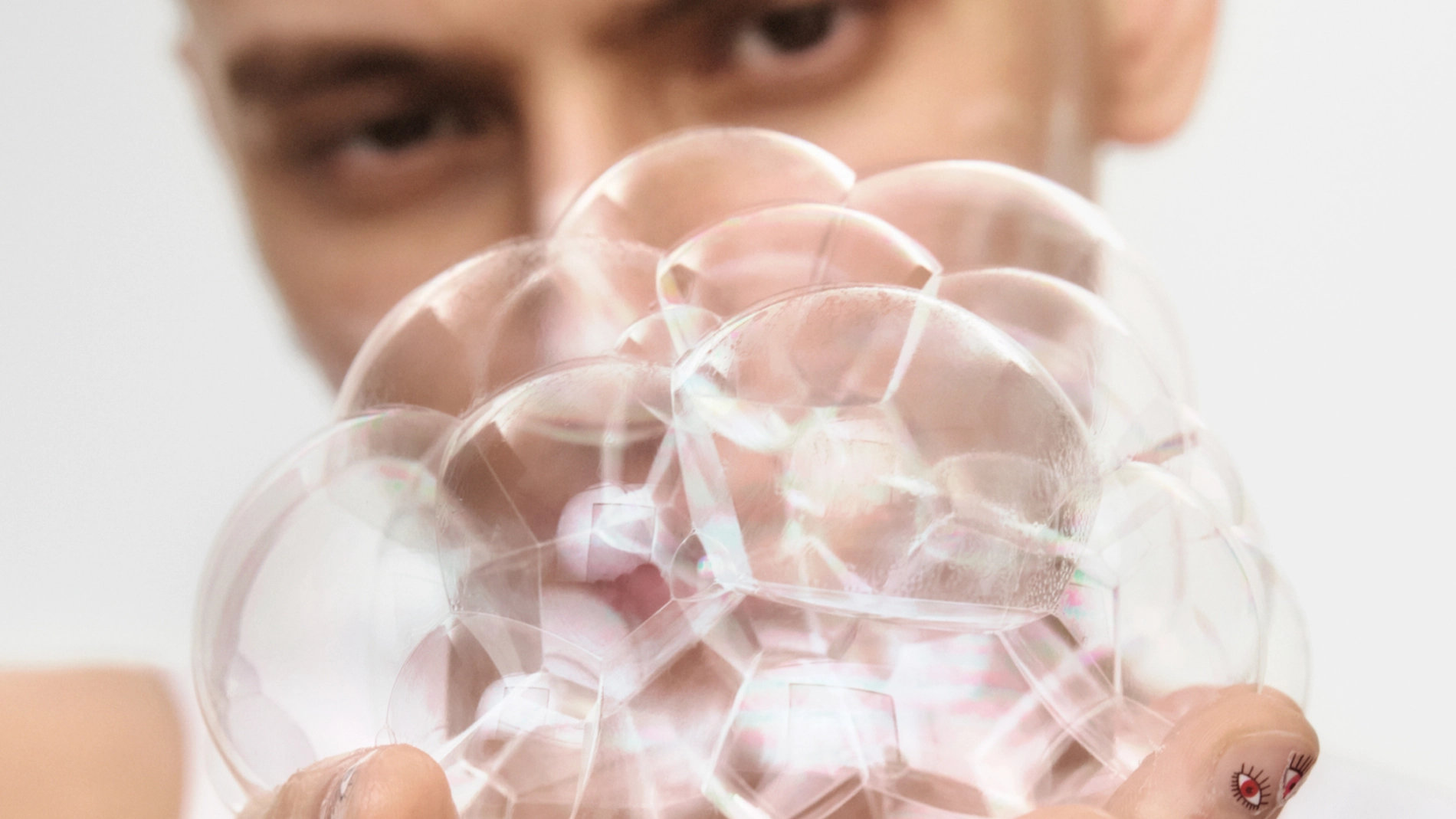 Närbild av en hand som håller en klunga genomskinliga såpbubblor framför ett ansikte i bakgrunden mot ljus bakgrund. Fokus ligger på de skimrande bubblorna och deras reflektioner.