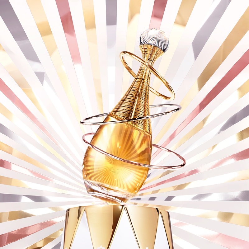 En gyllene Dior-flaska omgiven av metallringar, med en bakgrund av strålande linjer i guld, rosa och vitt.
