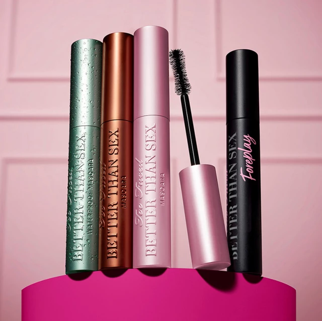 Fire mascaraer fra Too Faced er stilt opp på en rosa flate mot en rosa bakgrunn. Fra venstre mot høyre: en grønn tube, en kobberfarget tube, en rosa tube med børsten utenpå og en svart tube.