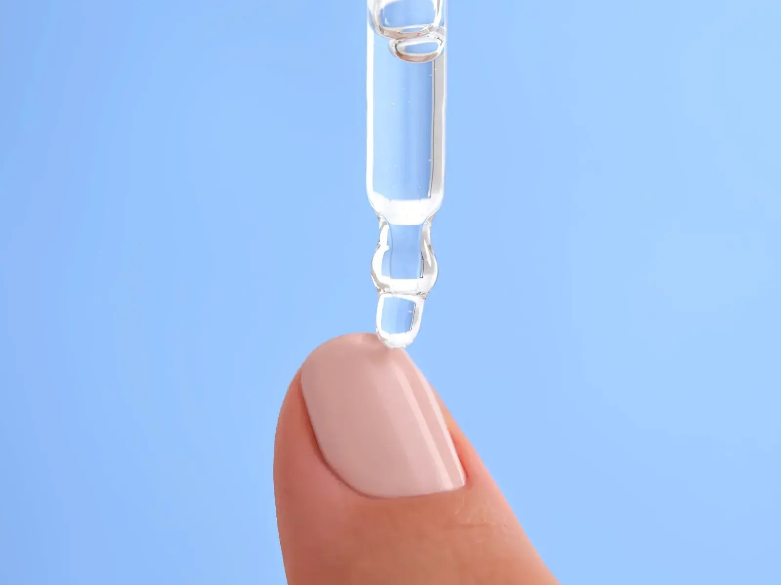 Nærbilde av en dråpe gjennomsiktig serum som påføres en velmanikyrert fingertupp mot en lys blå bakgrunn. Serumet dryppes fra en pipette, og fingertuppen har en lys rosa neglelakk.