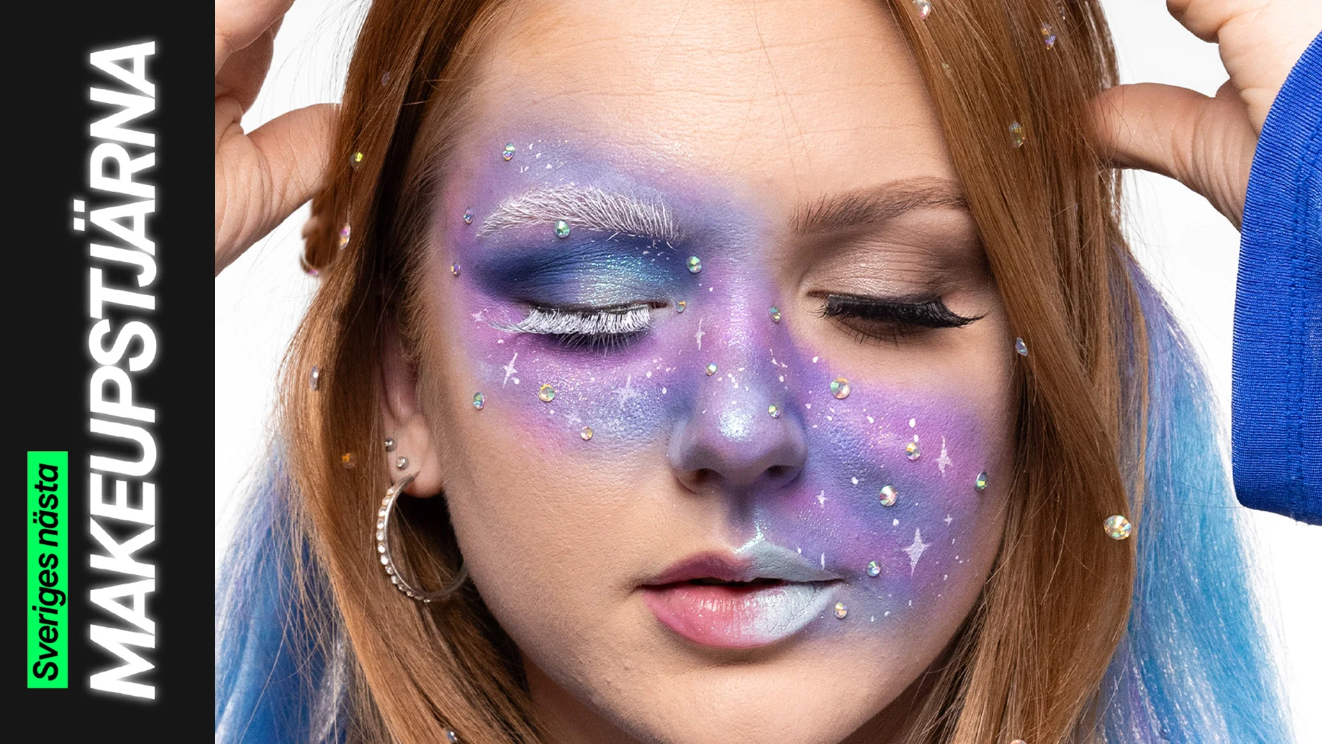 Närbild på galaxinspirerad makeup i lila, blå och silver med vita fransar, glittrande stenar och målade stjärnformationer, medan en ögonlockssida är öppnad och den andra stängd för att visa effekten.