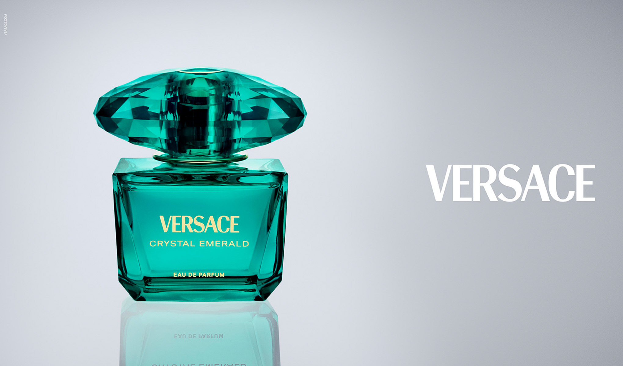 Versace Crystal Emerald-parfyme er avbildet i en elegant, smaragdgrønn flaske med en fasettert topp. Flasken står mot en lys bakgrunn, og Versace-logoen er synlig på høyre side.