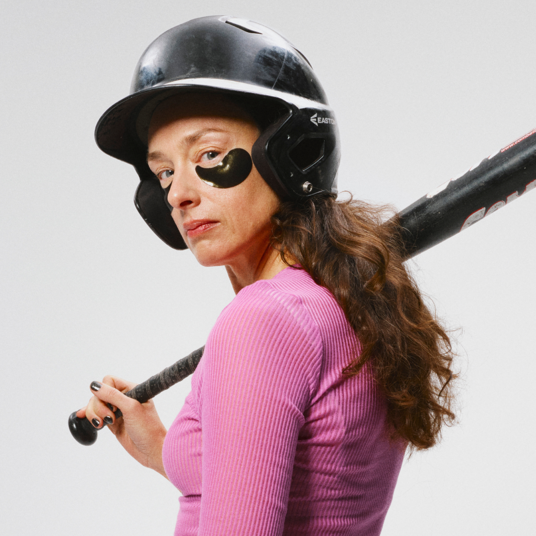 En person i rosa genser har på seg en svart hjelm og holder et baseballbat over skulderen. 