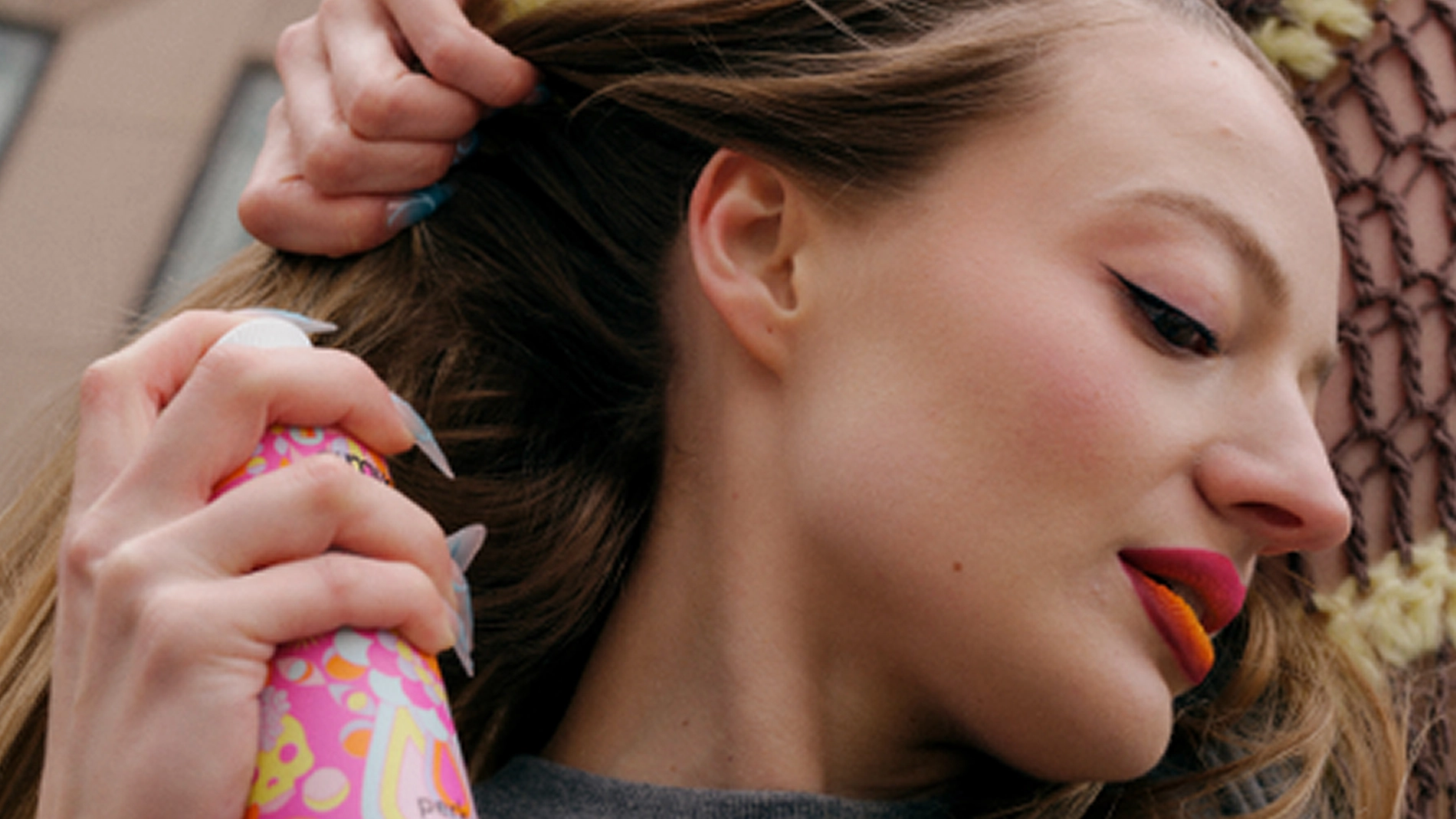 Person applicerar hårspray från en färgglad sprayflaska i håret, med utsläppt hår och makeup med eyeliner och färgstark läppstift.