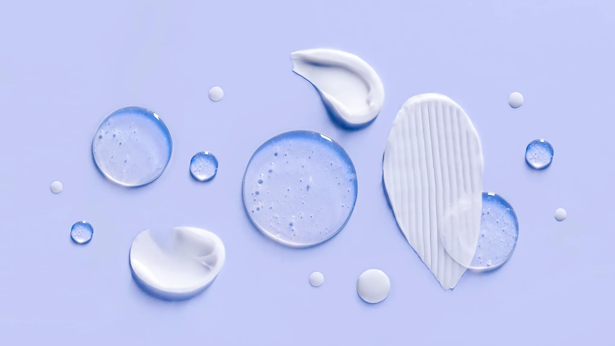 Ulike teksturer av hudpleieprodukter – kremer, geler og serum – spredt utover en lys lilla bakgrunn.